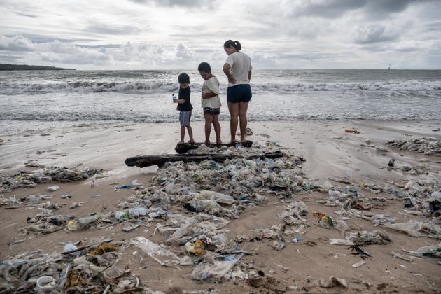 A woman with two boys stand on some wood which is covered by plastic trash at a beach in Bali.